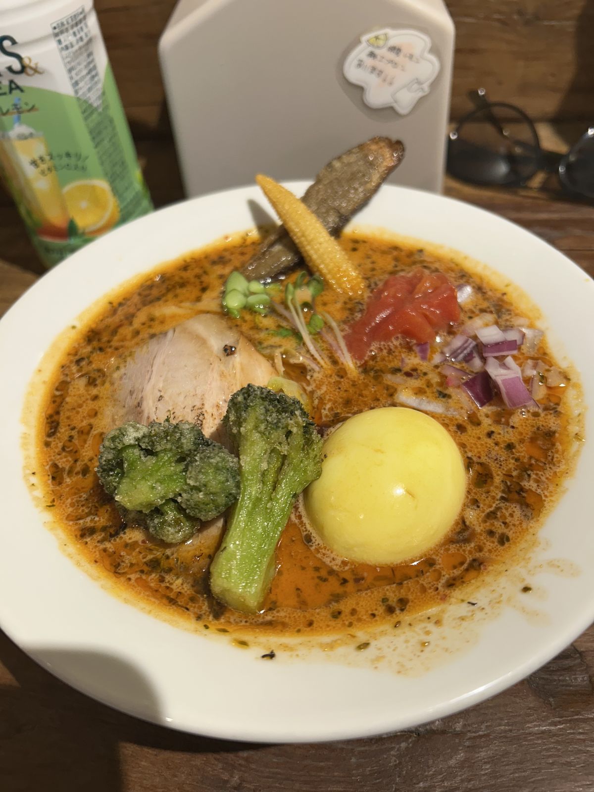 Bol de ramen épicé au bouillon orangé, avec une tranche de poulet, un œuf entier, du brocoli, un épi de maïs miniature, des dés de tomate et d'oignon rouge