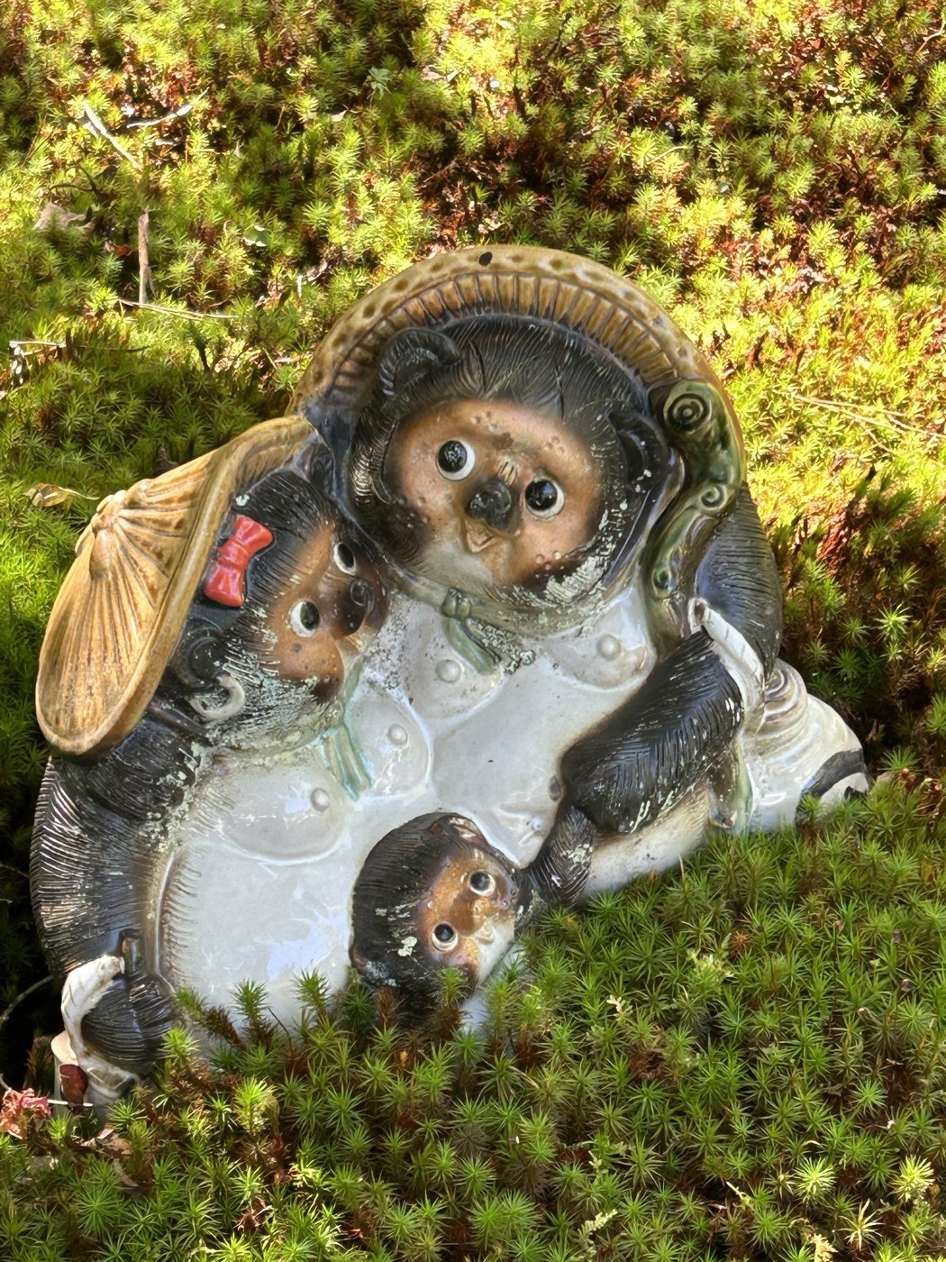 Statuette en céramique d'une famille de tanuki (parent, enfant au ruban rouge, bébé) posée dans la mousse