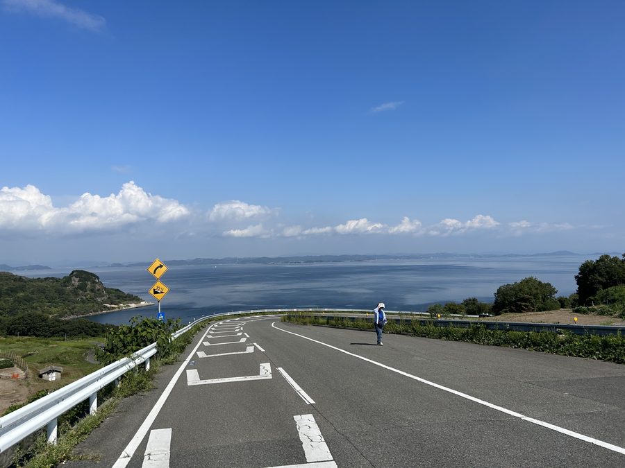 A coastal road running down to the Seto Inland Sea on the island of Teshima, with a cyclist stopped on the tarmac.