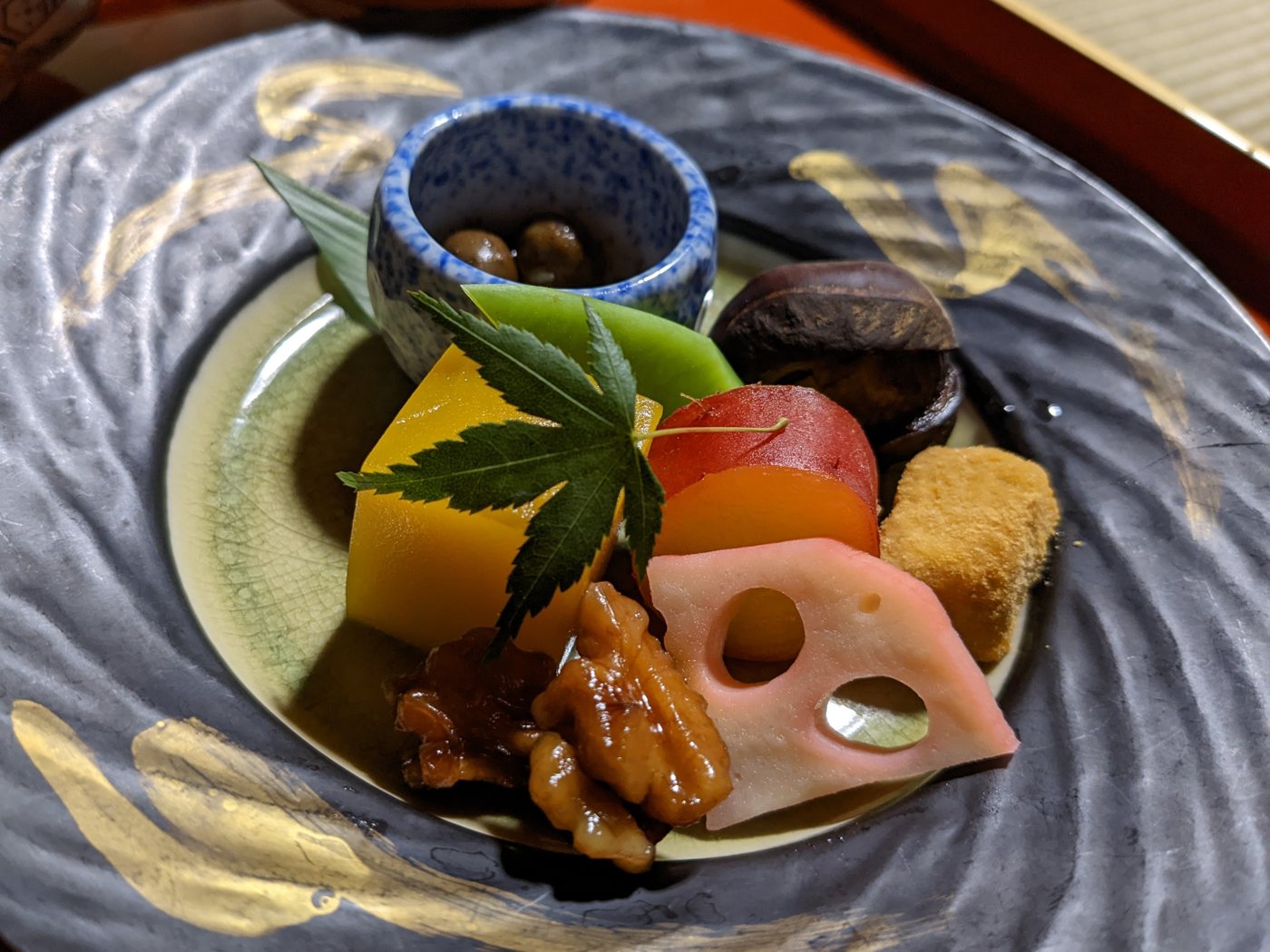 Une assiette de laque grise portant sept petites préparations, dont une racine de lotus, une patate douce et des haricots noirs, sous une feuille d'érable