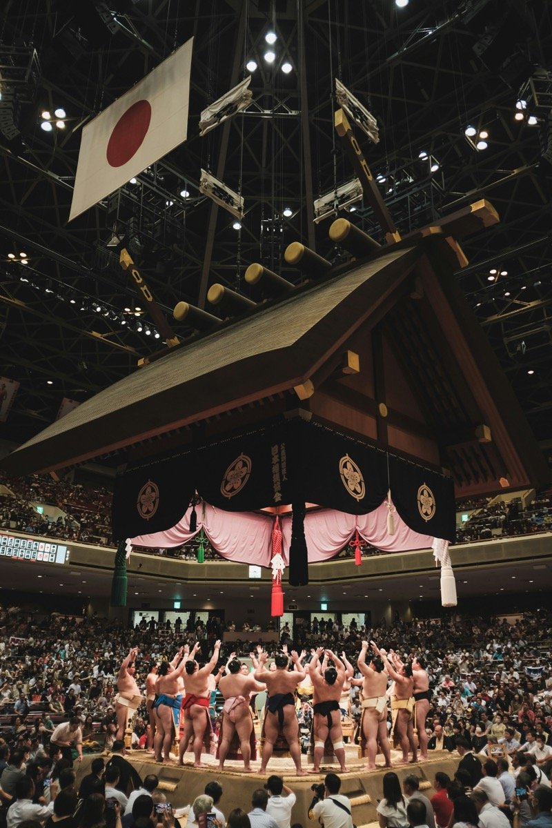 Vue plongeante sur le dohyō de l'arène de Ryogoku pendant le dohyō-iri, le cortège de rikishi qui lève les bras devant la foule, sous le toit suspendu et le drapeau japonais