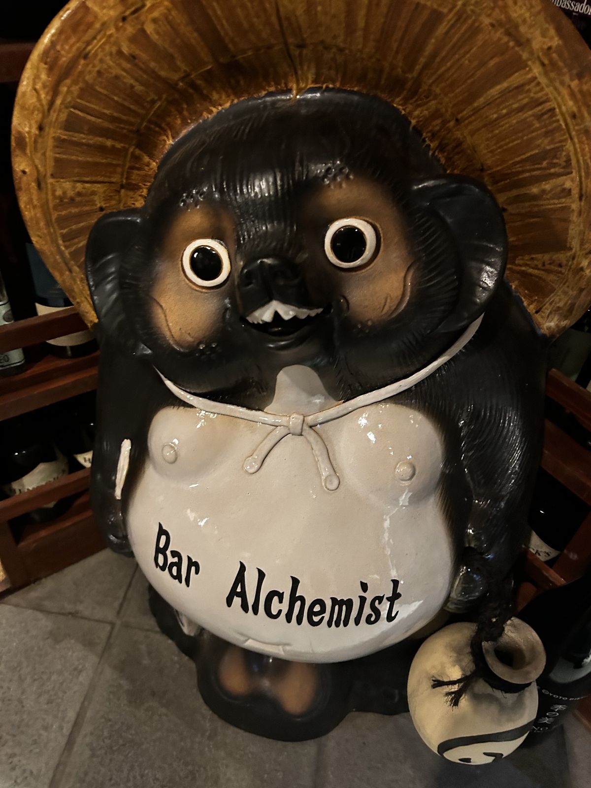 Statue de tanuki en céramique noire et blanche, chapeau de paille, flasque de saké à la main, « Bar Alchemist » peint sur le ventre