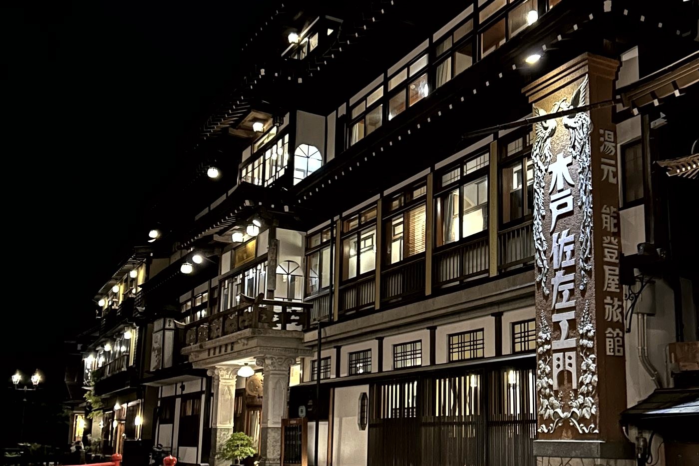 The lit wooden facade of a ryokan at Ginzan Onsen at night, with a carved sign on the right.