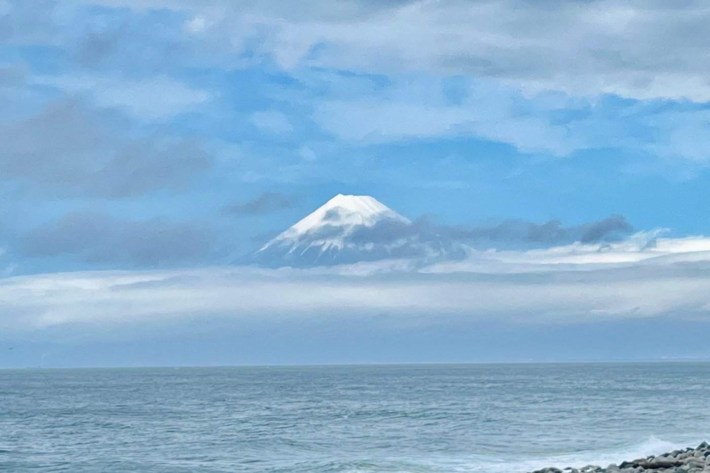 Le mont Fuji enneigé vu depuis une plage de galets, mer et nuages en arrière-plan.