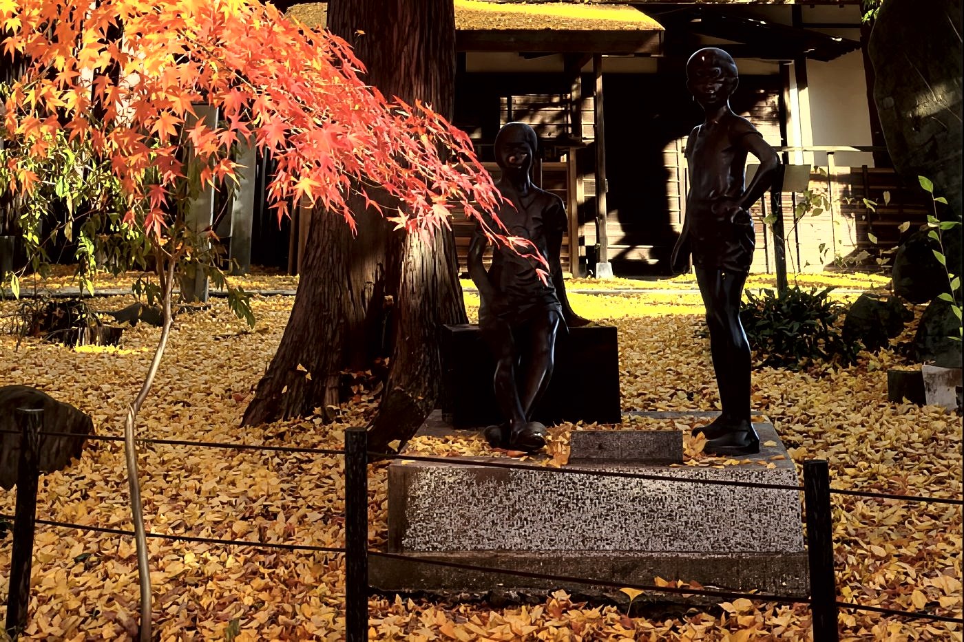 Statues de bronze de deux enfants sous un érable, devant une maison au toit de chaume couvert de feuilles de ginkgo dorées.