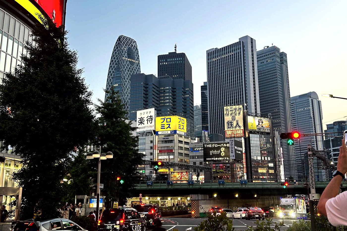 Carrefour de Nishi-Shinjuku au crépuscule, la tour Cocoon (Mode Gakuen) au loin, néons et circulation.