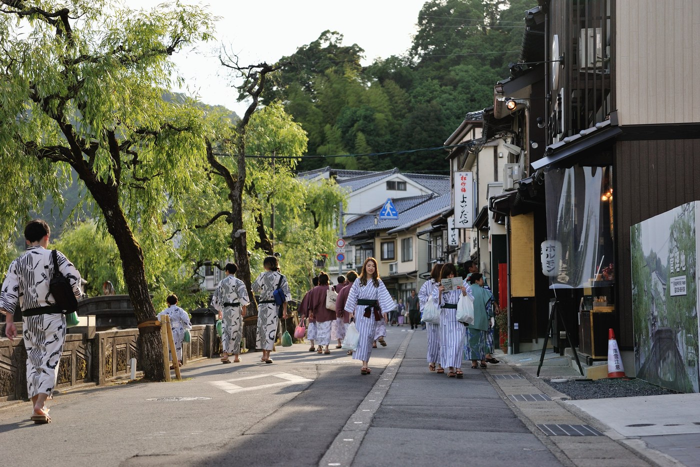 Visitors in yukata walking the willow-lined canal at Kinosaki Onsen as night falls.