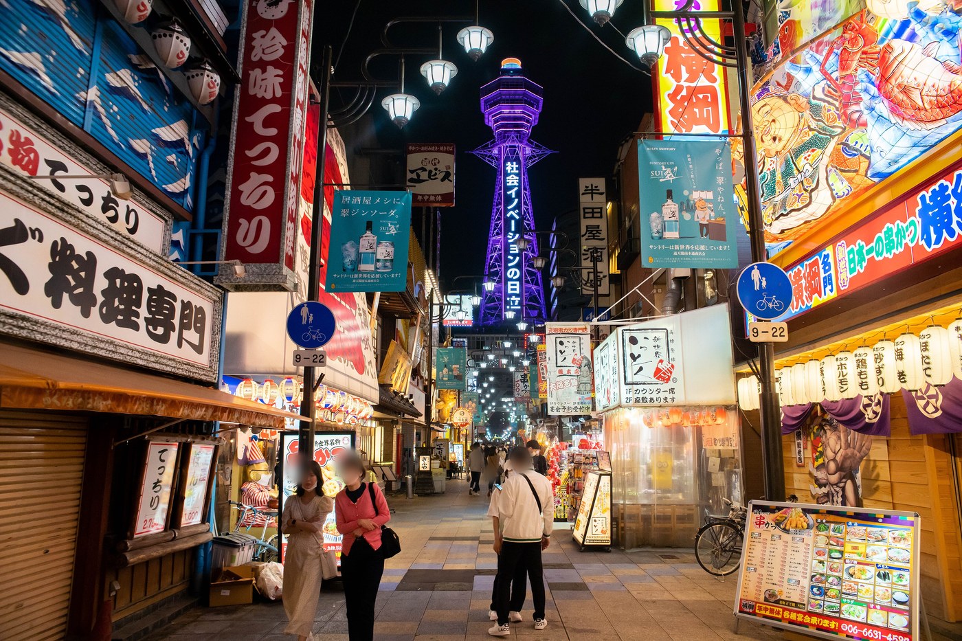 The Tsutenkaku tower lit purple at night, seen from a busy lane in the Shinsekai district.